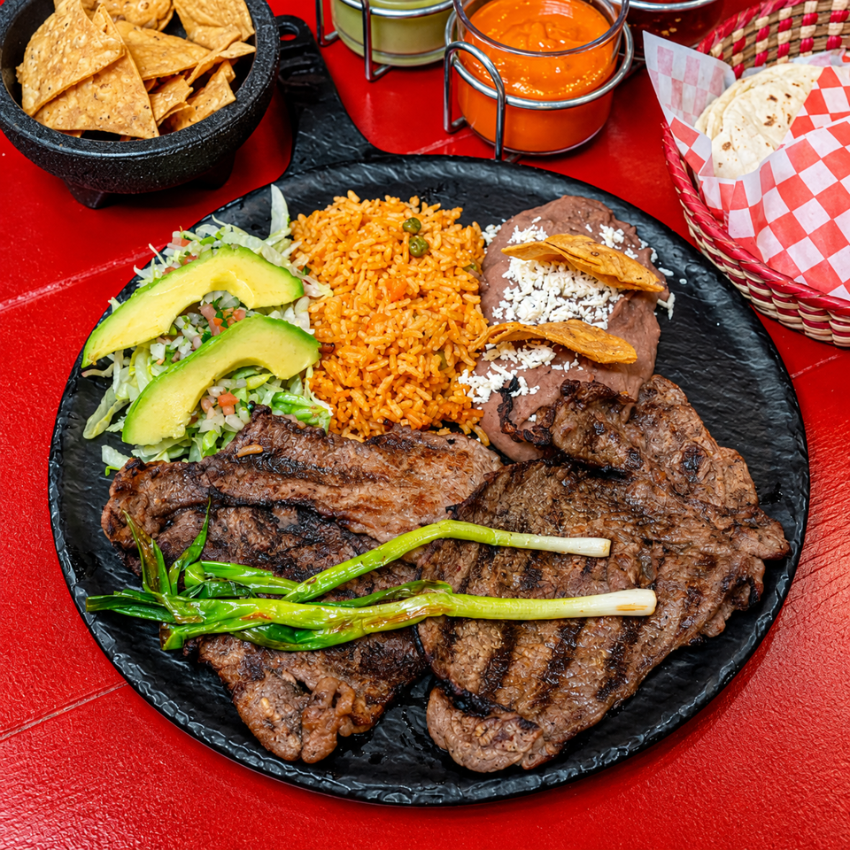 Mexican steak platter with grilled beef, rice, refried beans, avocado salad, tortillas, chips, and salsa on a red table.