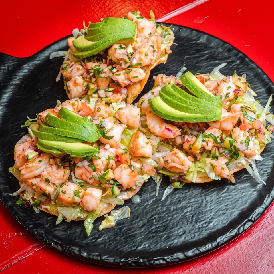 Three shrimp tostadas topped with avocado, lettuce, diced onion, tomatoes, and herbs on a black plate.